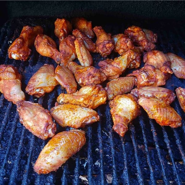 Many seasoned chicken wings and drumettes cooking on a dark grill grate.