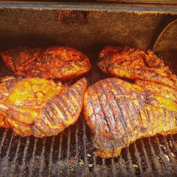 Four large pieces of seasoned meat, possibly pork or chicken, grilling on a dark, smoky barbecue.