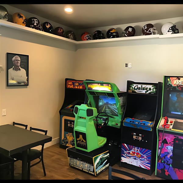 Arcade games, a table with chairs, and football helmets on a shelf in a game room.