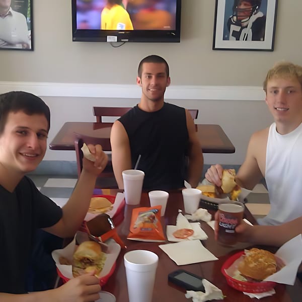 Three young men enjoying Fast Eddie's sandwiches and drinks at a table with a TV in the background.