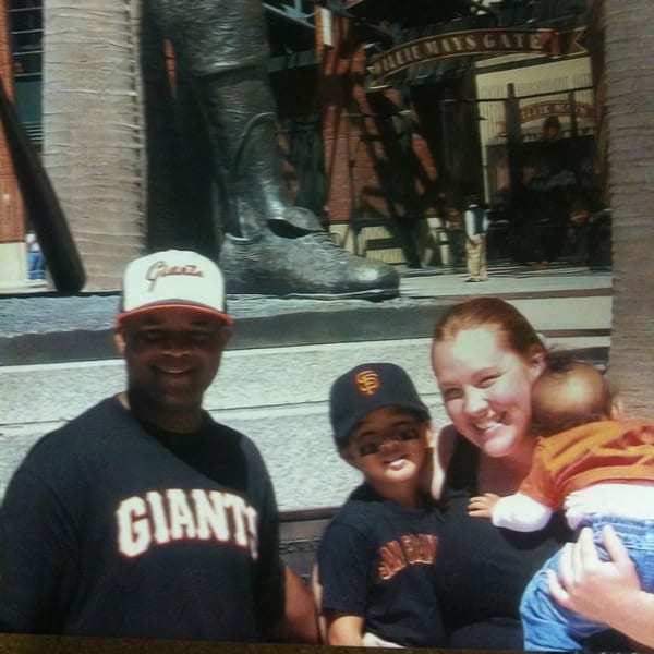 A family of four, including a baby, smiling in front of a large statue and "Willie Mays Gate" sign.
